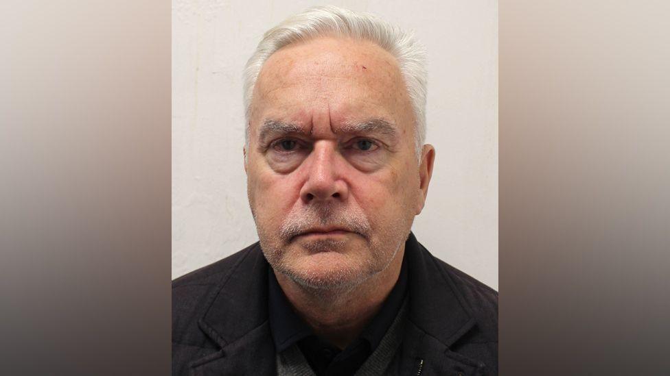 A custody image of a man with white hair and stubble on his face. He is wearing a black jacket and looking directly at the camera with a serious expression.