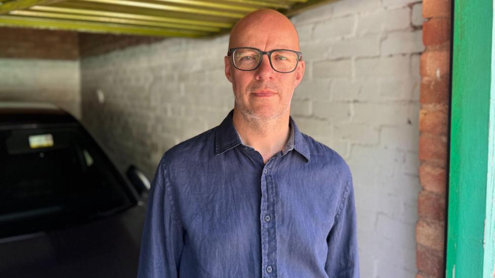 A bald man wearing glasses, wearing a dark blue shirt, stood in a garage with a car behind him.