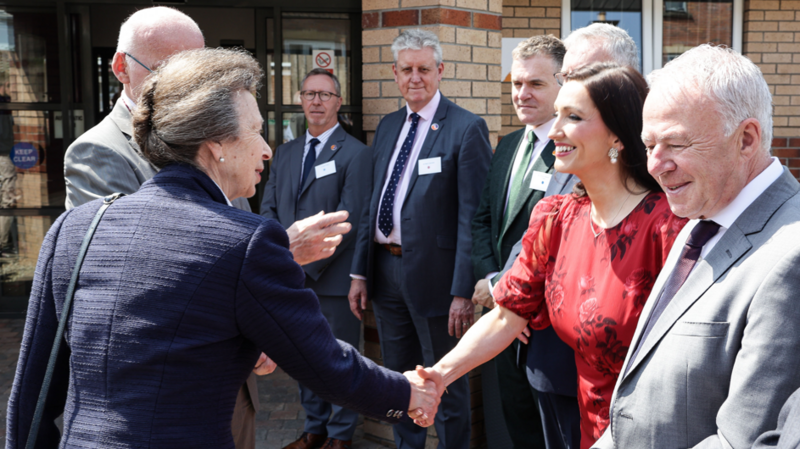 The Princess Royal, a woman with grey hair and wearing a blue jacket, greets Emma Little-Pengelly, a woman in a red dress, who has dark hair. There are other men with grey hair in the photo.