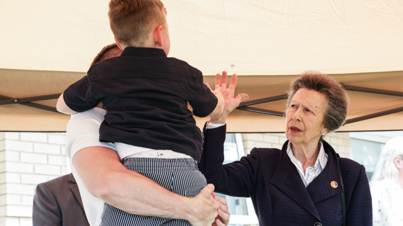 The Princess Royal, a woman with grey hair, wearing a blue coat, puts her hand up to a boy in a black top, being held by a man with a white top.