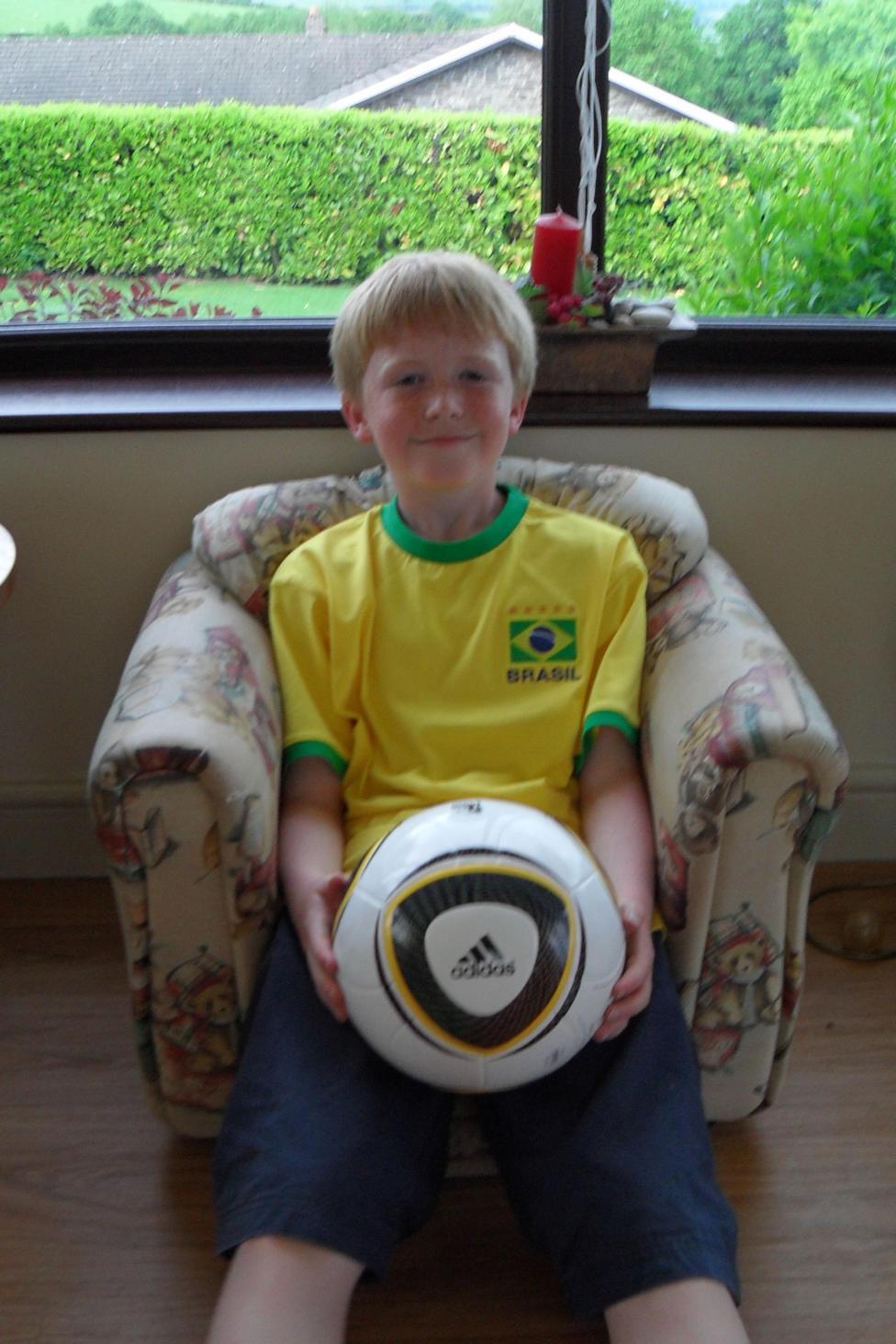 10-year-old Ieuan sits cantered in a padded armchair, holding a full-size white Adidas football in his lap with both hands. He has short blond hair. The ball features bold black and gold curved panels and the Adidas logo clearly visible at the front, making it the focal point of the image. The child is wearing a bright yellow Brazil football shirt with green trim and a crest reading “Brasil,” visually linking the clothing to the sport