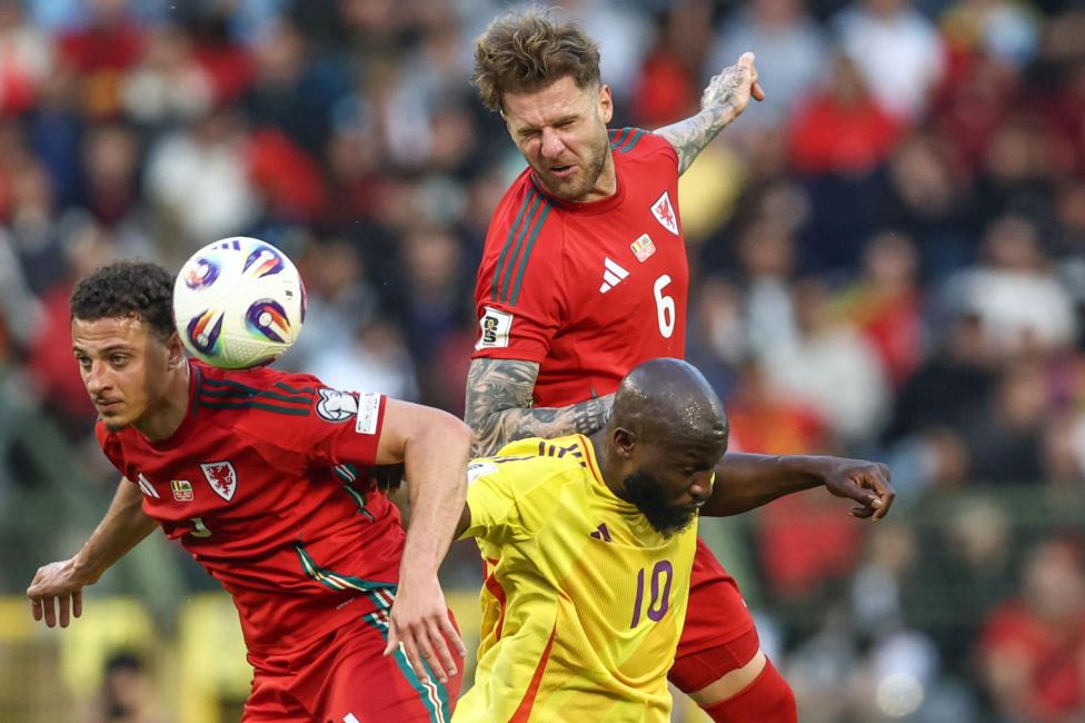 Wales footballers Joe Rodon and Ethan Ampadu are either side of Belgium's Romelu Lukaka as they challenge for the ball. Rodon has leapt in the air to head the ball and has his eyes closed while leaning on the back of Lukaku