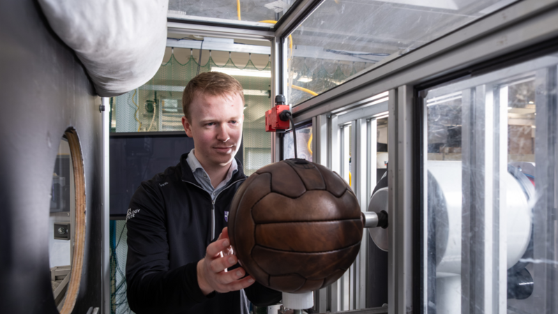 Phillips holding a brown leather ball and placing it into a metal contraption. He has blonde hair and is wearing a black jumper. 