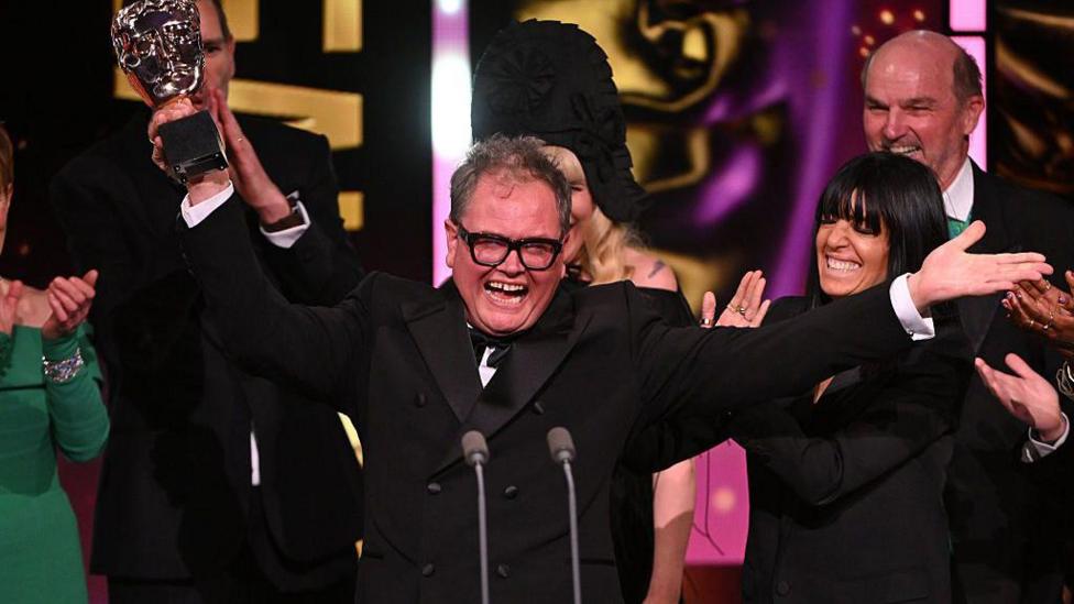 Comedian Alan Carr in a dark suit with his trademark large glasses holds a Bafta award in one hand with his arms opened wide in celebration. Claudia Winkleman is beside him.