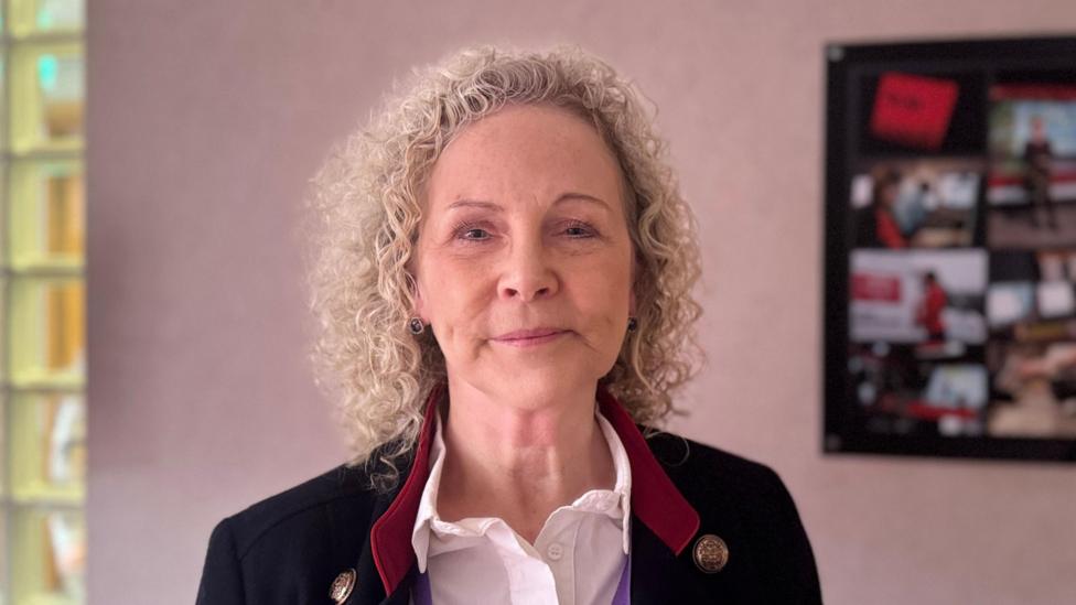 Dr Jacqui Montgomery-Devlin. She is an older woman with curly, blonde hair. She is looking at the camera with a neutral expression on her face. She is stood in a room with a pale pink wall, and a framed photo. She is wearing a white shirt, with a black blazer and red collar.