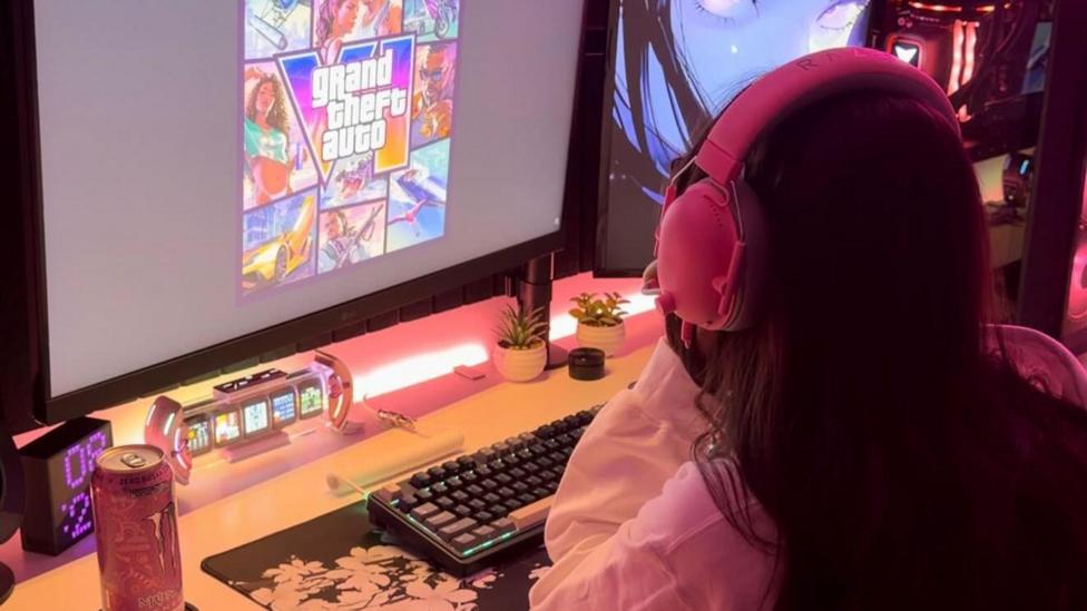 A behind the shoulder view of a woman - the content creator Alogirlx - staring at her computer screen which has the artwork and logo for GTA 6 on it. She is wearing pink headphones and her desk with a keyboard is lit up brightly with pink LED lights
