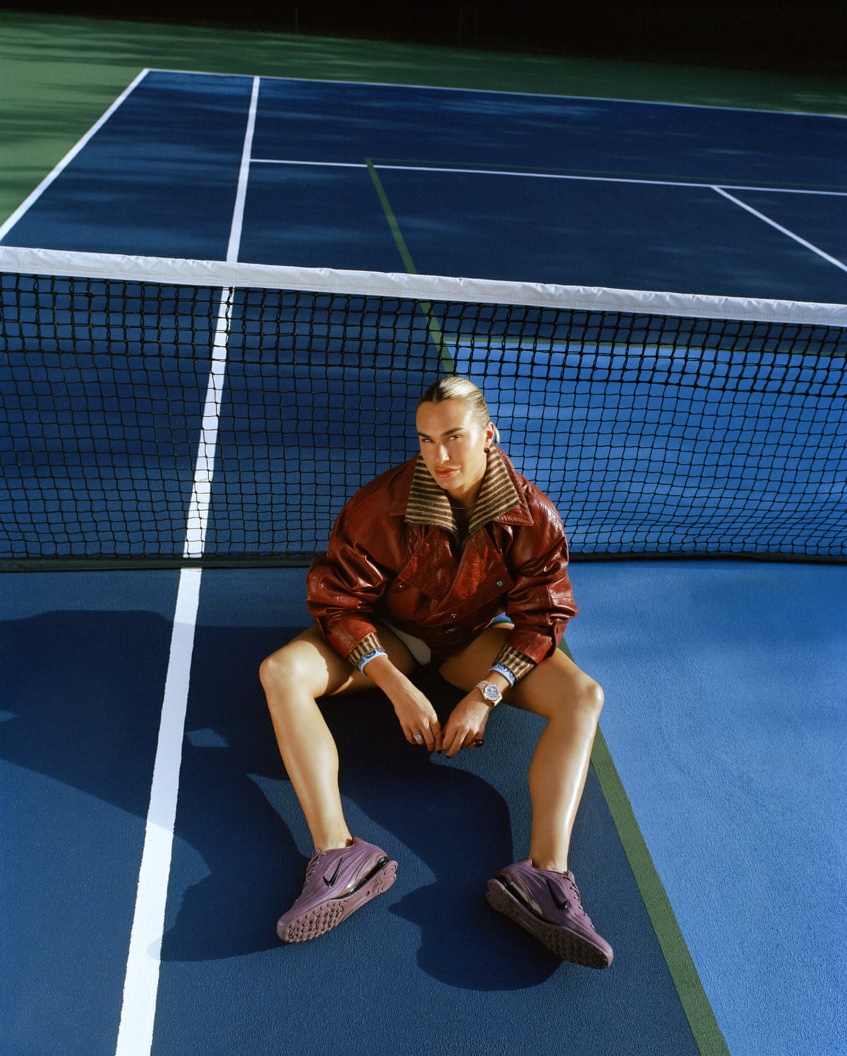 Sabalenka sitting on a tennis court with her legs slightly bent and out in front of her and her forearms on her thighs, wearing a brown jacket, shorts and purple trainers