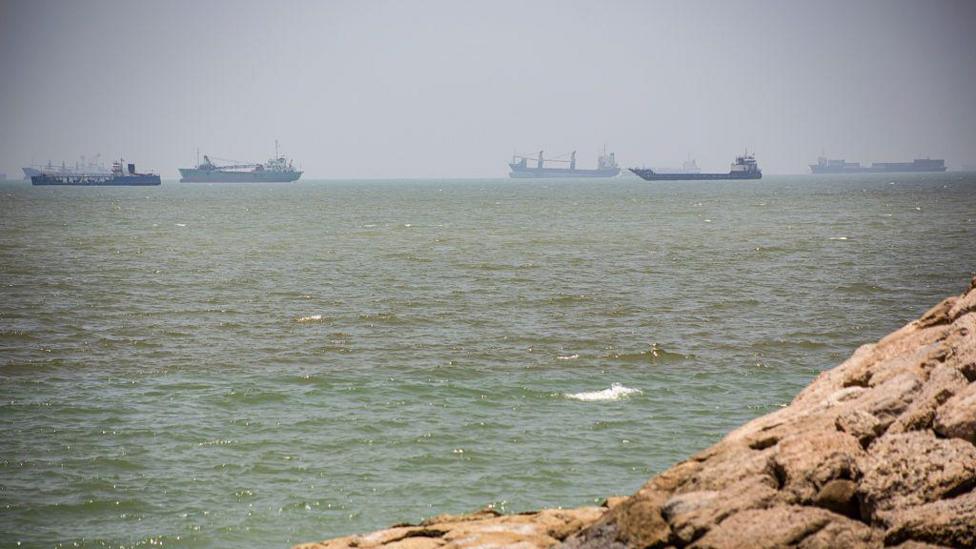 The strait of Hormuz pictured from rocks placed in the right corner of the image. It looks out onto water, in the distance are five vessels.