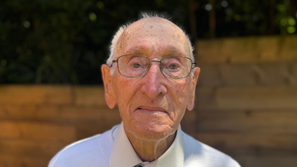 A head and shoulders image of an elderly man wearing a white shirt and glasses. He is looking directly into the camera.