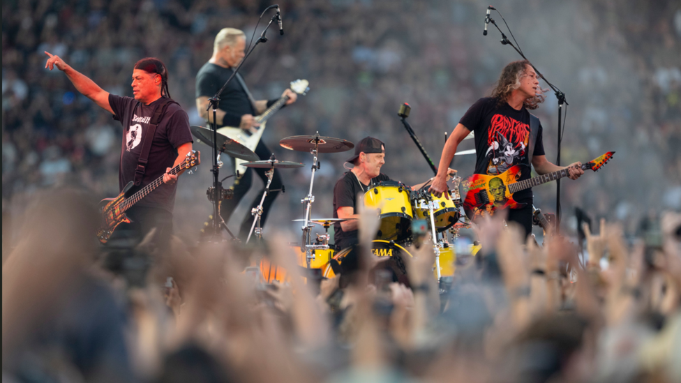 The band play on stage. Raised hands can be seen in the foreground, with seats full of fans seen behind them. A guitarist raises his hand in the air, with the other band members behind him. 
