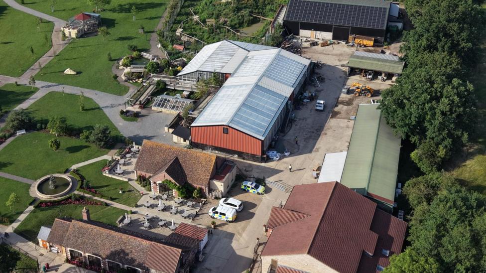 An aerial drone shot of the whole site showing several medium and large buildings or farm sheds, with three police cars parked in the courtyard. There are also formal gardens on the left side and lots of greenery.