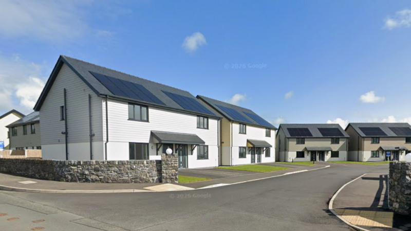 A cul-de-sac of semi-detached modern homes lining a road. They all have solar panels on the roof with wooden cladding on the top of the buildings, and each has a grey porch roof. 