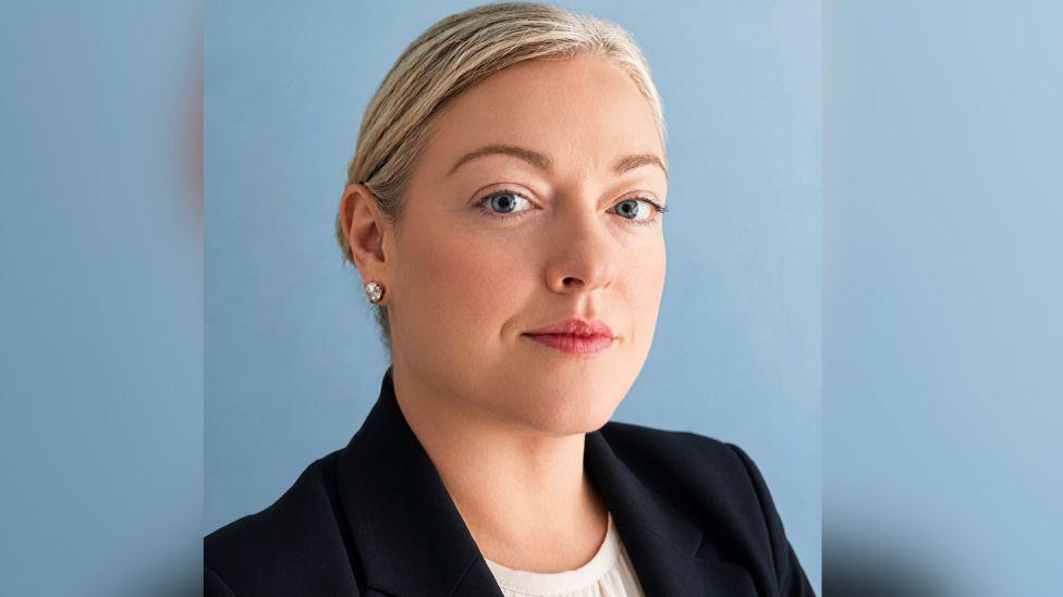 A blonde woman looking at the camera against a blue background. She is wearing a dark jacket 