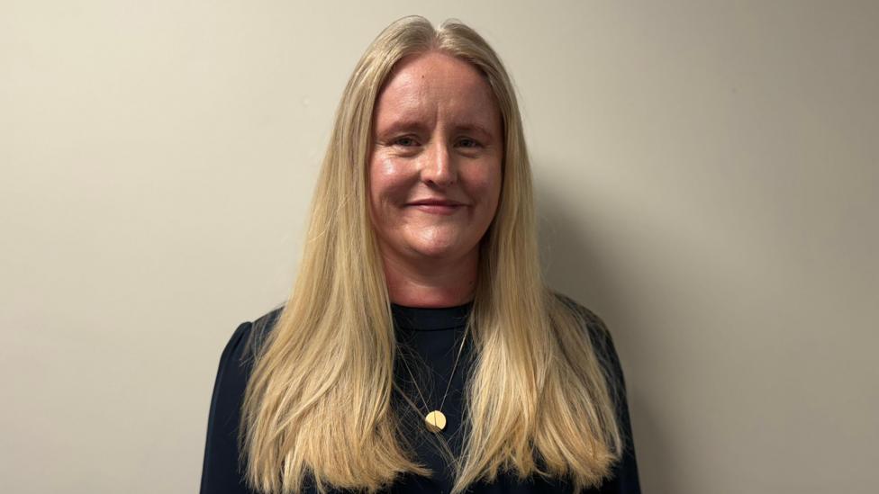 A woman with long blonde hair stands looking at the camera, she is wearing a black jumper and a gold necklace. 