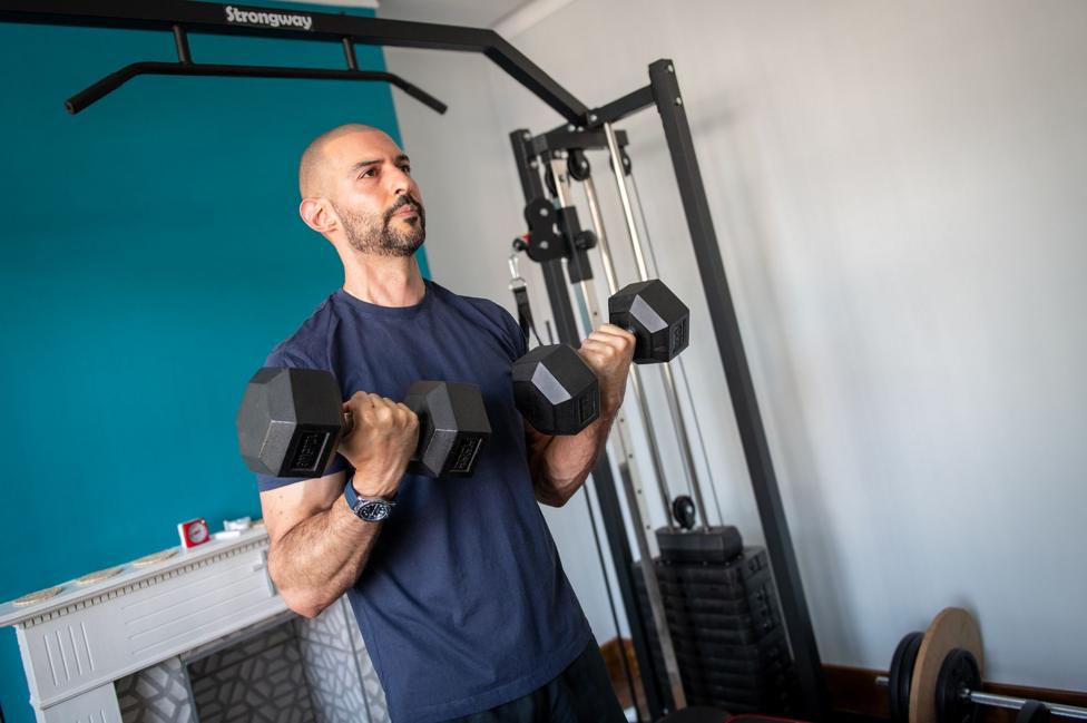 Marios in a dark blue t-shit and shorts doing bicep curls with two dumbbells 