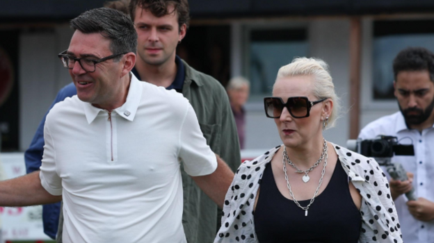 Andy Burnham wears a white polo top with a zip and black glasses. He is walking with his wife Marie-France van Heel, and has a hand on her back. Van Heel has light blonde hair and wears large black sunglasses, thick silver jewellery and a black vest with an open white shirt which has black polka dots on it.