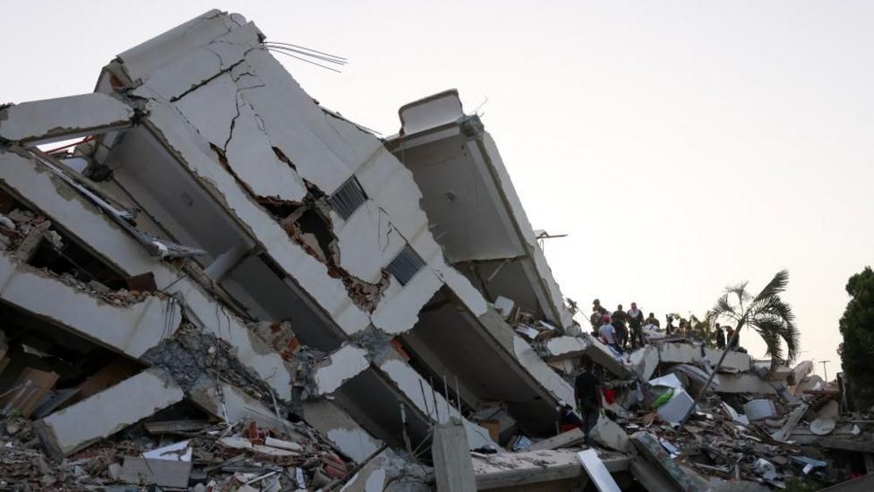 A large, crumbled building with a group of people climbing onto it.