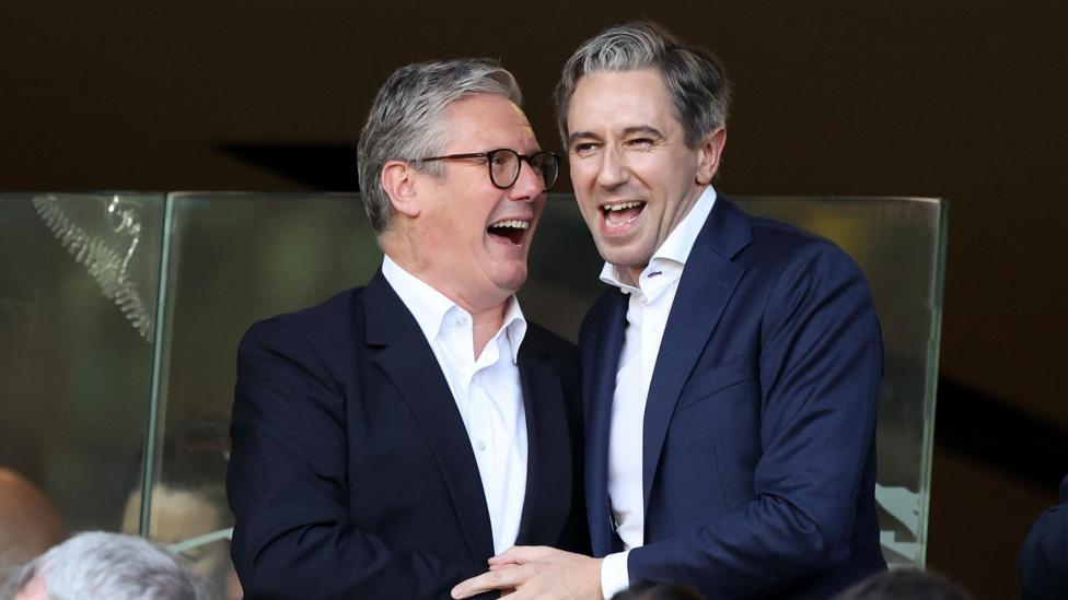 Sir Keir Starmer and Simon Harris. They are both smiling in celebration. Starmer has grey hair, wearing a dark blazer and white shirt. Harris has grey hair, wearing a navy blazer and white shirt.
