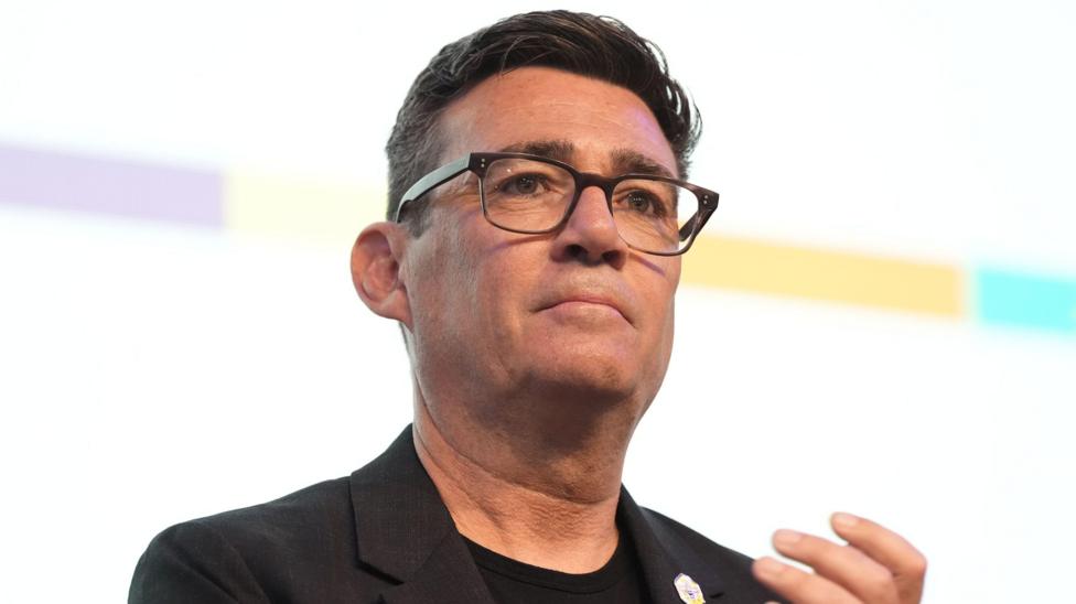 Andy Burnham. He has dark hair, wearing glasses, a black blazer and black t-shirt.
