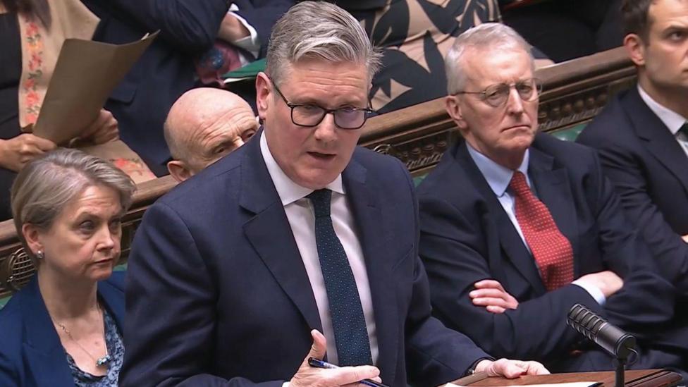 Keir Starmer speaking in the House of Commons. He has grey hair, wearing glasses, a navy blazer, white shirt and navy tie. Other MPs are behind him.