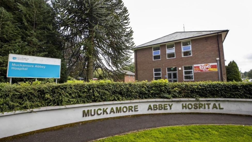 The exterior of Muckamore Abbey Hospital. There is a sign featuring the name of the hospital and it is also written on a low concrete wall. There is a hedge above this wall and a patch of grass in front. Behind it you can see two red brick buildings.