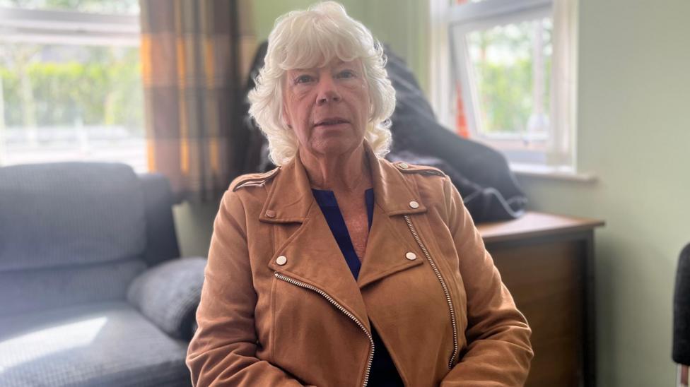 A woman with grey hair and a brown jacket. She is sitting down, with a grey sofa behind her.