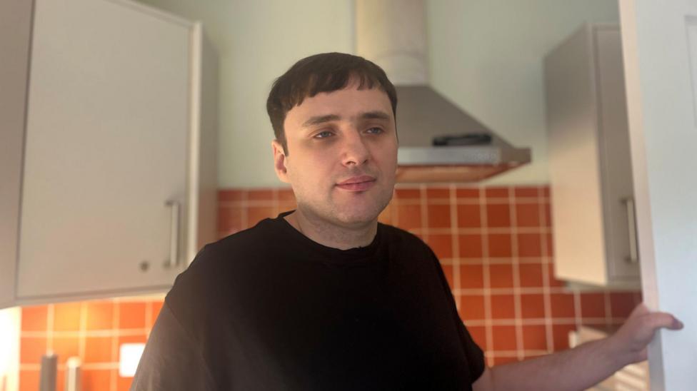 Mid-shot of Timothy Jones standing in a kitchen, his left hand holding onto a white door. He has brown hair and a black t-shirt.