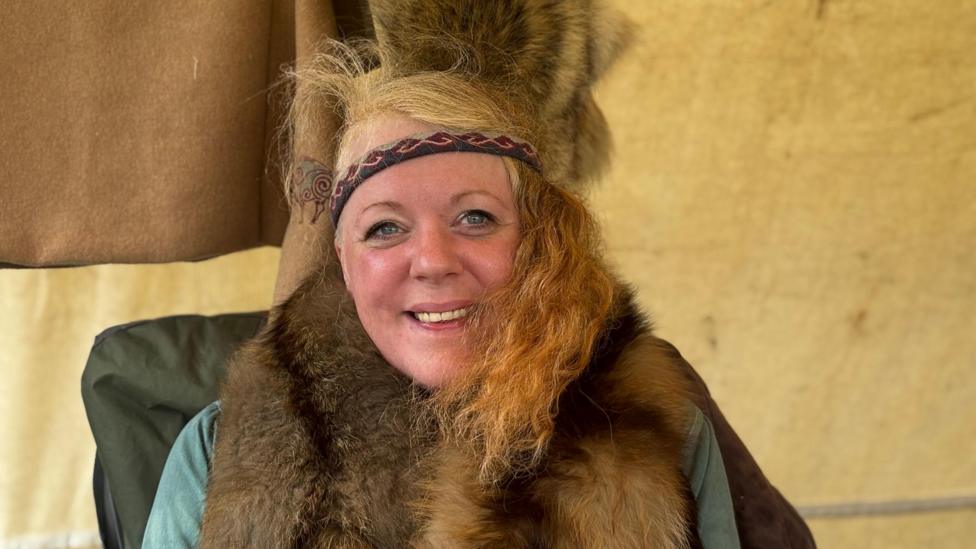 A woman with blonde hair, wearing a blue top with a fur scarf. She is also wearing a headband. 