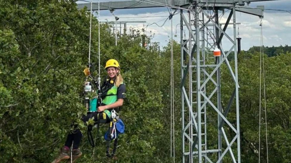 A woman in a harness hanging from a pylon next to a tree canopy. She has long blonde hair and wears a yellow hard hat, green vest and black t-shirt and trousers, with brown walking boots.