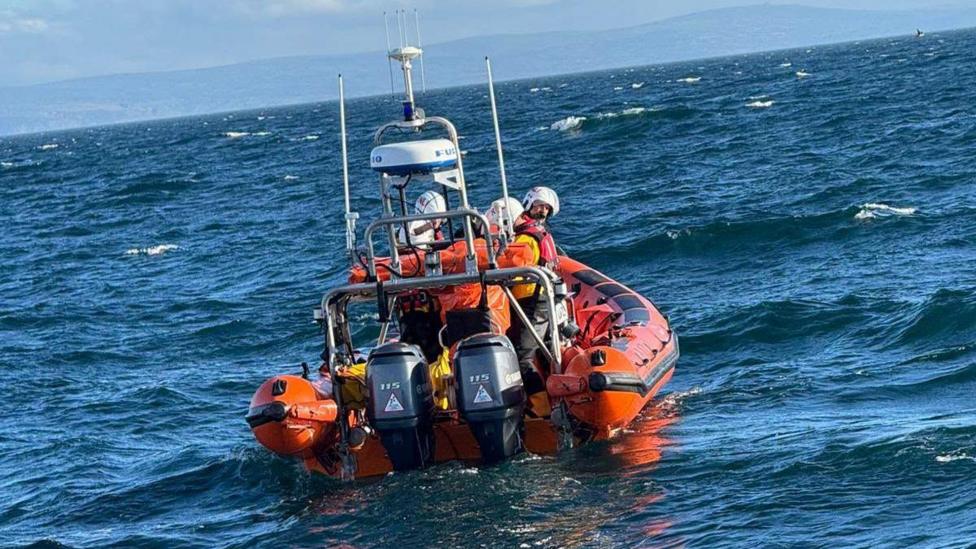 Orange life boat with crew wearing helmets on board seen in the sea from behind with engines at the back