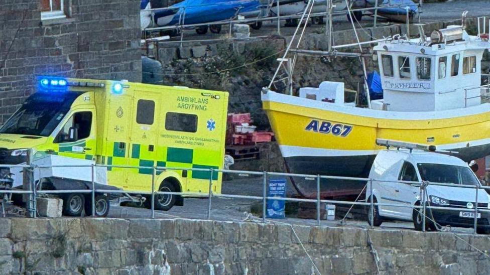 A pier with a docked yellow and black boat, and a yellow ambulance parked up next to it.