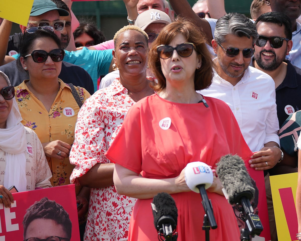 Lucy Powell speaks at an event: she is wearing a bright red dress with elbow-length loose sleeves and dark sunglasses. She stands in front of a crowd in the sunshine. 