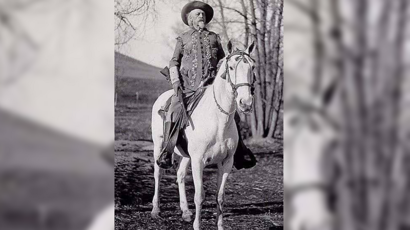 Sitting on a white horse, an older Cody is pictured with white hair wearing and ornately decorated jacket and cowboy hat, carrying a gun in his right hand 