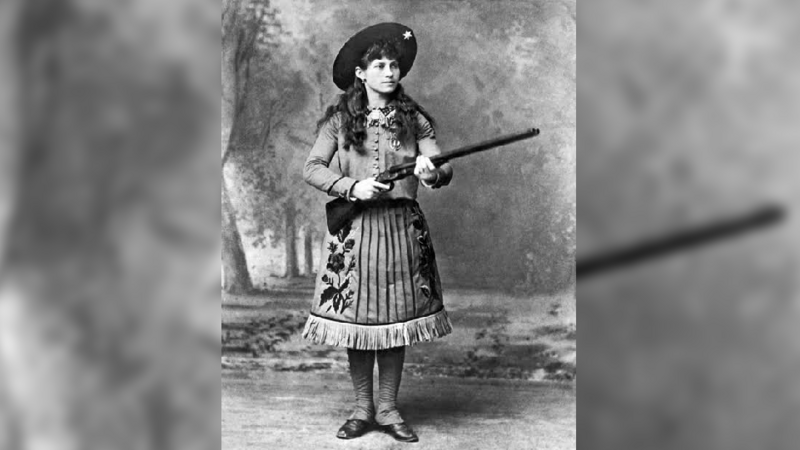Oakley, shown with long brown curly hair beneath a wide-brimmed hat, is posing with a rifle