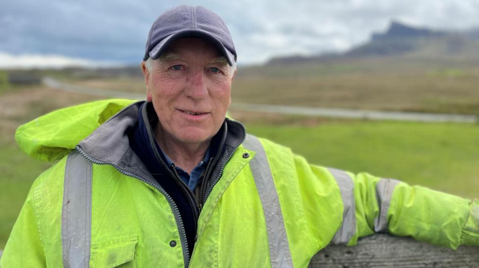 Calum Beaton is outdoors with moor and hills behind him. He is wearing a blue cap and a yellow high-visibility jacket over a fleece and a shirt.