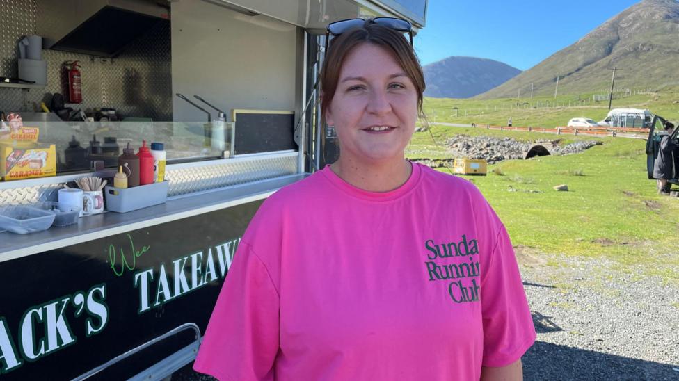 Sarah MacKinnon is standing outside her outdoor catering trailer. She has her brown hair tied up and is wearing a bright pink T-shirt, and a pair of sunglasses on top of her head. It is a bright sunny day with hills in the background.