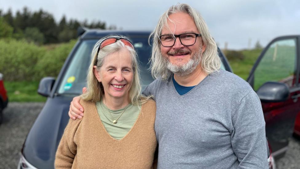 Beate and Heiner Delbach are standing outside in front of a car. Heiner who has long grey hair, a beard and is wearing glasses and a long-sleeved top, has an arm around Beate. Beate also has long grey hair, has sunglasses on top of her head and is wearing a brown jumper over a green top.