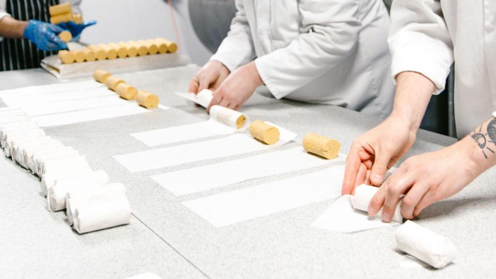This image shows the hands of three people rolling cylindrical pats of butter into paper wrappers.