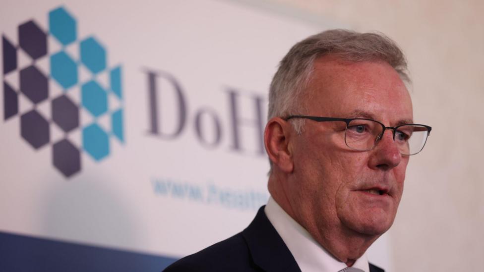 Health Minister Mike Nesbitt speaking to media at the Department of Health, Castle Buildings, Stormont Estate, Belfast. He has short grey hair, wearing glasses, a dark blazer and white shirt. Department of Health branding is behind him.
