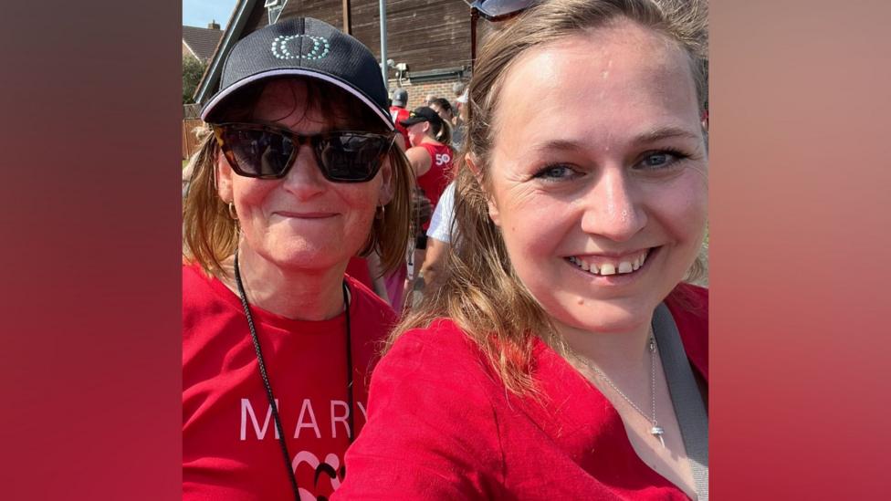 Two women both wearing red; the woman on the left is wearing sunglasses and a sun hat. She has short brown hair and smiling at the camera. Another woman has blonde hair and is also wearing a red top and a small silver necklace. They are both smiling at the camera 