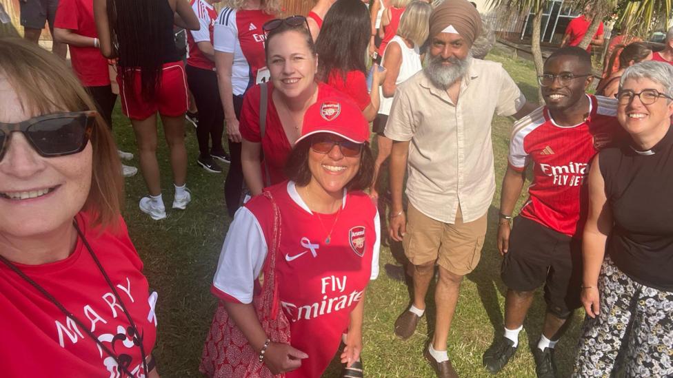 A woman wearing black sunglasses on the left of the photo while she takes a photo. Also in the frame is three woman and two men. One woman is wearing a red top and a red sunhat, while another woman behind her is smiling and wearing a red blouse. Next to her is an eldery man wearing a turban, a buttoned up shirt, long shorts and loafers. Next to him is another man wearing a red shirt and black shorts. There is a woman at the end of the photo on the right side and she is wearing floral trousers and a black top. They are standing outside on the grass and there are a lot of people outside behind them. 