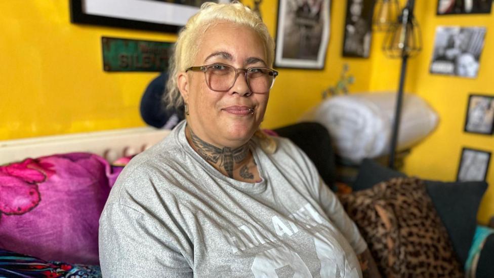 Dee is sat in her living room on a sofa, the wall behind her is yellow and there are framed pictures on the walls. She is smiling at the camera and wearing a grey printed t-shirt and glasses.