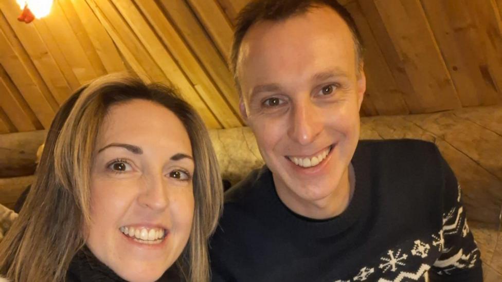 A man and a woman sitting inside a wooden cabin. They are shown from the shoulders up and are both smiling at the camera. The woman, on the right has brown shoulder length hair and is wearing a black polo neck jumper and the man has short brown hair and is wearing a nordic-style black and white jumper. 