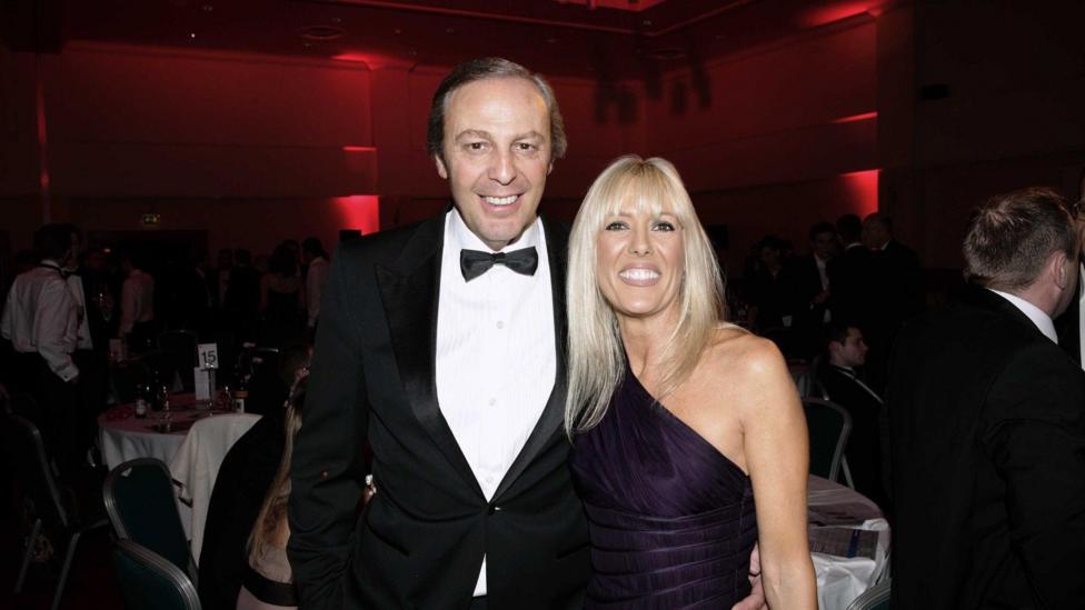 A man in and woman in black tie and formal dress standing arm in arm smiling at the camera