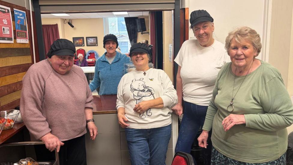 Five women are standing next to a kitchen hatch smiling at the camera.