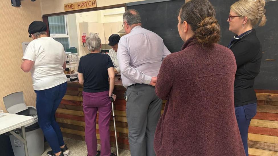 Five people stand in line at a kitchen hatch with the sign 