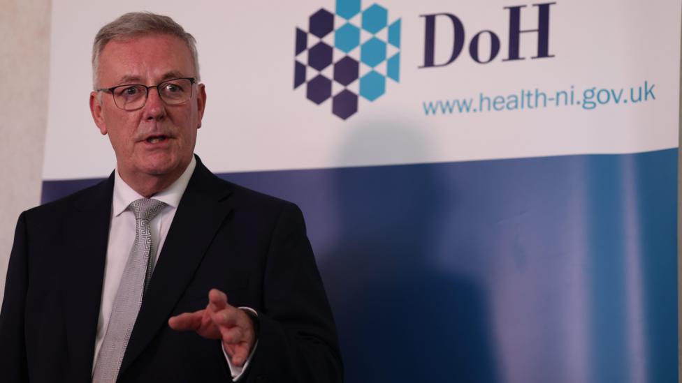 A man with white hair in a dark suit and glasses speaking in front of a white and blue DoH banner.