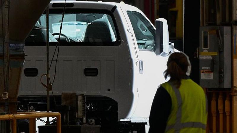 A Ford truck on a production line, passing through inspection checks, at a plant in Ohio. A female worker wearing a high-vis jacket looks on. 
