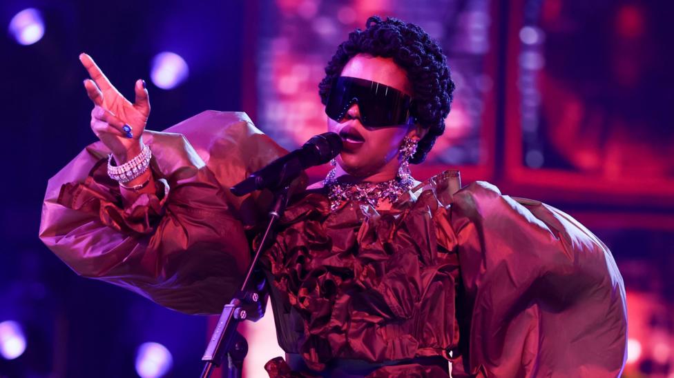 Lauryn Hill in large dark glasses and an elaborate fress with balloon sleeves, singing into a microphone and gesturing with one raised hand