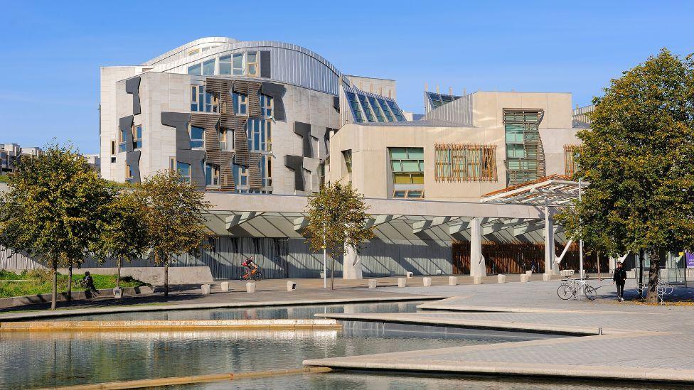 A general view of the Scottish Parliament on a sunny day, with a manmade pond in the foreground. The stone building has wood panelling on the outside. 